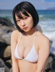 A woman in a white bikini sitting on a rock by the ocean.