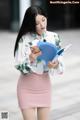 A woman in a pink skirt holding a blue folder and a cell phone.